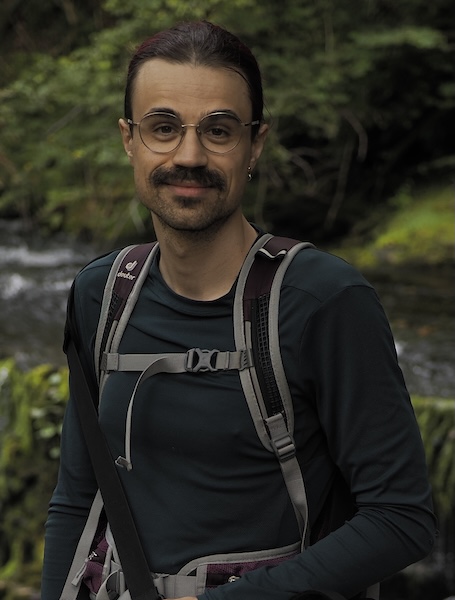 a person, troy, stands in front of a waterfall. they are wearing a green top and have many straps across their chest from a rucksack and camera bag. their hair is tied up and they have a moustache.
