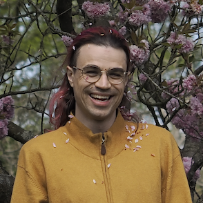 a person (troy) with pink hair and glasses smiles as pink petals fall from the cherry tree above across their face and onto their mustard-coloured cardigan